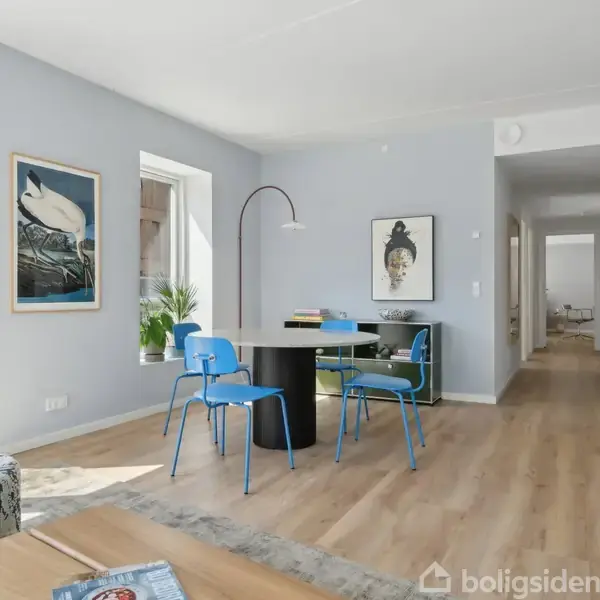 En lys stue med trægulv har et rundt bord omgivet af blå stole. En buet lampe hænger over bordet. Vægge prydes af kunstværker, og store vinduer lader lys strømme ind.