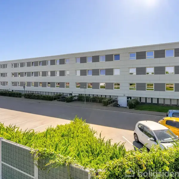 En lang, hvid boligblok står stille under en klar, blå himmel. I forgrunden er der parkerede biler langs en asfalteret vej. Grønt buskads grænser vejen.