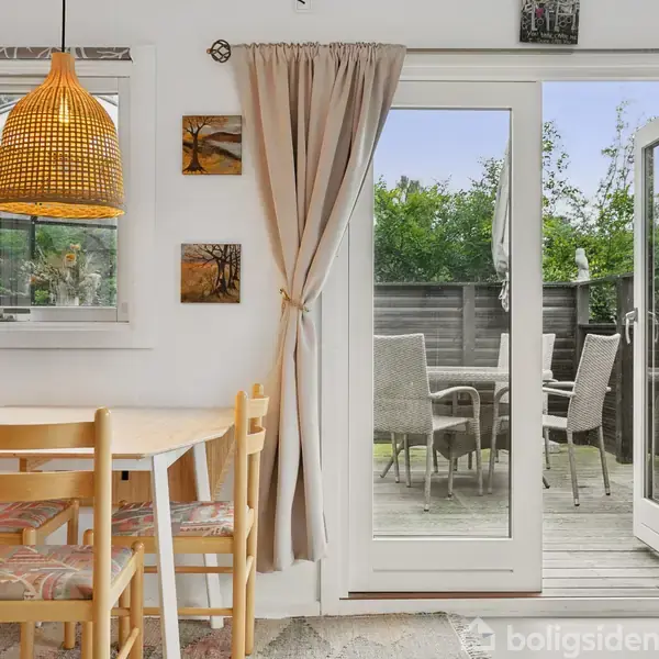 Et spisebord med fire stole står ved et vindue med en hængende lampeskærm. Dørene åbner ud til en terrasse med havemøbler og grønne planter i baggrunden.