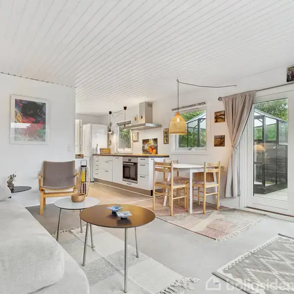En lys stue med en grå sofa og runde sofaborde i et åbent køkken-alrum. Bord og stole står ved et vindue omgivet af grønne planter udenfor. Tepper ligger på gulvet.