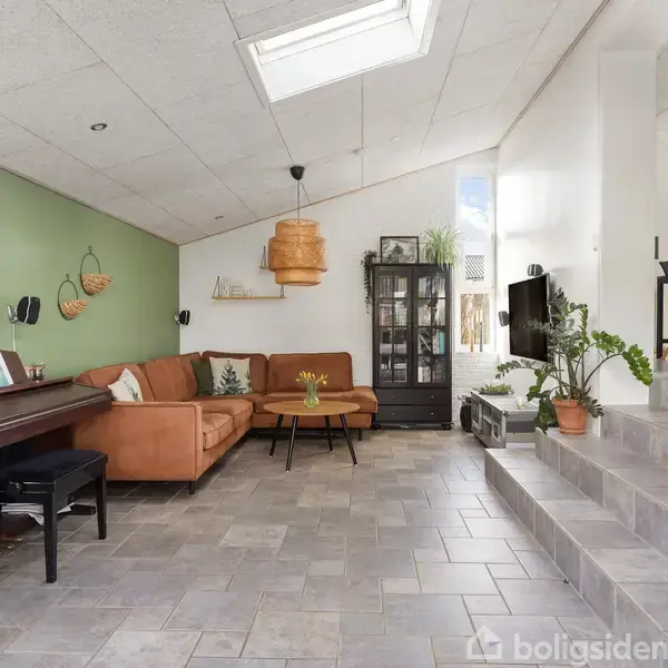 En stue med brun sofa og sofabord. Et klaver står ved en grøn væg med maleri. Et vitrineskab og en lampe er i nærheden. Trin fører til spiseområde. Ingen tekst fundet.