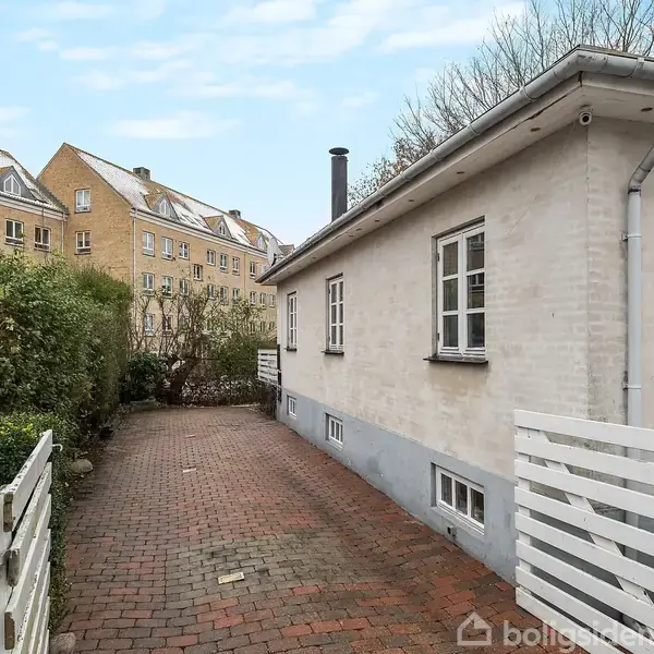 Et hvidt hus med flere vinduer står på en belagt sti i et bymiljø. Omkring huset er hække og flere større bygninger kan ses i baggrunden.