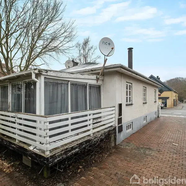 Hvide hus med udestue og satellitantenne, står på en brolagt indkørsel. Omgivet af buske og træer, ses andre huse i baggrunden, på en rolig villavej.