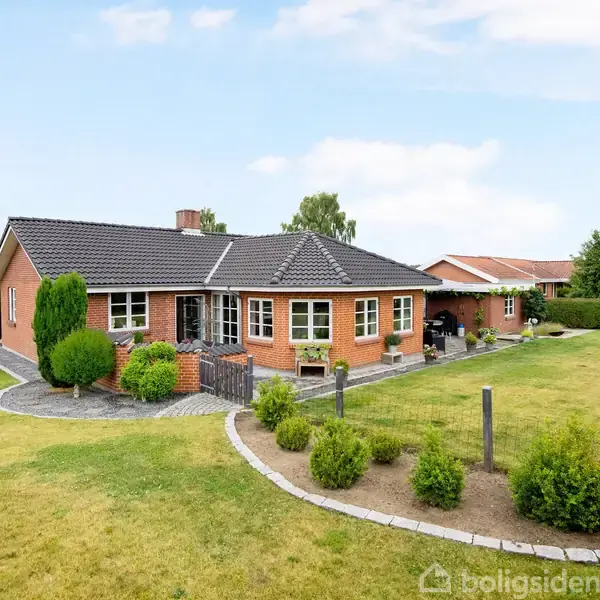 Rødt murstenshus står centralt med velplejet have omkring. Græsplæner og buske omkranser huset, mens himlen er let overskyet. Fladt tag og indkørsel ses i baggrunden.