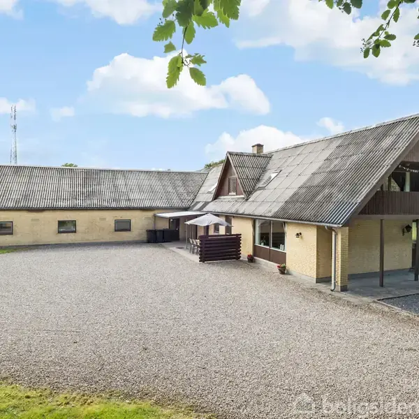 Et L-formet landhus med gul facade og mørkt tag står på en grusplads omgivet af grønne områder, under en blå himmel med enkelte skyer.