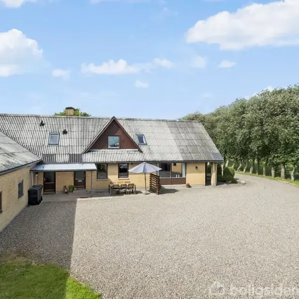 En gårdbygning med gult murværk og en gruset gårdsplads; en parasol og havemøbler står foran. Omkring er der grønne træer og en blå, let skyet himmel.