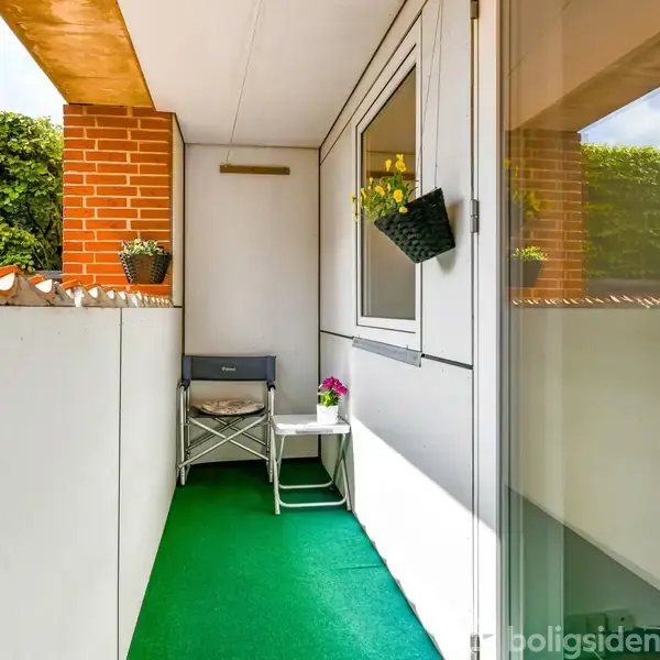 Balkon med grøn tæppebelægning, en stol og et lille bord med blomster, flankeret af hvidmalede og murstensvægge. Blomsterkasser hænger på væggen, omgivet af en hæk udenfor.