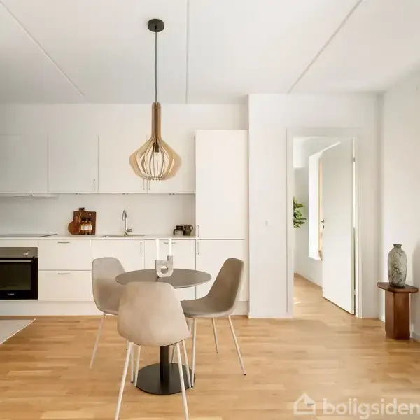 Spisebord med fire stole står i moderne køkken med hvide skabe og træelementer. En stor lampe hænger over bordet. Vindue til højre giver lys til rummet.