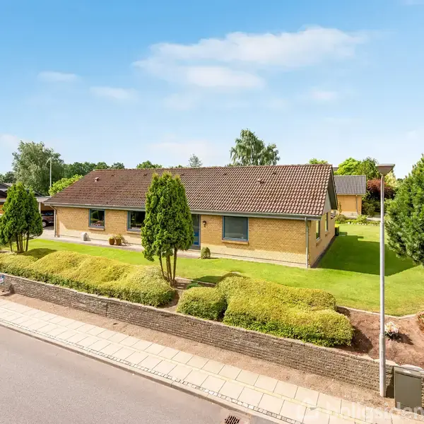 En gulstensbungalow med brun tag ligger i en pænt anlagt have omkranset af en lav mur. Huset har en velplejet græsplæne og buske, omgivet af en stille gade.