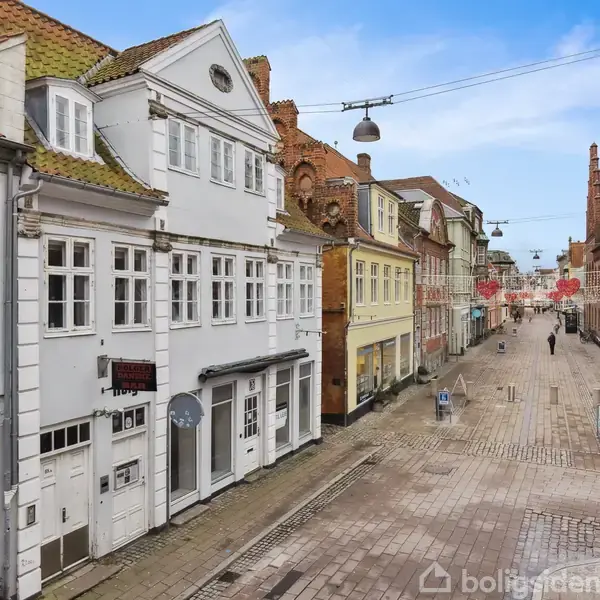 Gade med hvide og røde murstenshuse strækker sig fremad. Folk går afslappet. Juledekorationer hænger fra lygtepæle. Brosten dækker fortovet. Himlen er skyfri og blå.