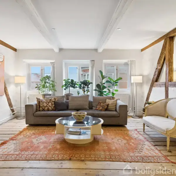 En sofa står centralt i en stue på et rustikt trægulv. Sofaen er flankeret af planter og lamper ved vinduerne. Kunst hænger på væggen, og en rund sofabord står foran.