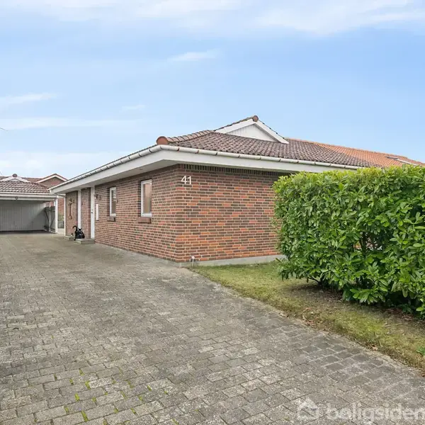 Murstenshus med nummeret 41 står på en brolagt indkørsel. Der er grønne hække på begge sider, og i baggrunden ses en garage. Himmel er blå med få skyer.