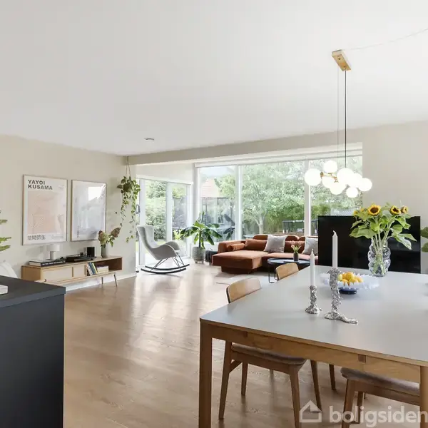 En lys stue med spisebord omgivet af stole. En lampe hænger over bordet. I baggrunden står en orange sofa nær store vinduer med udsigt til en grøn have.