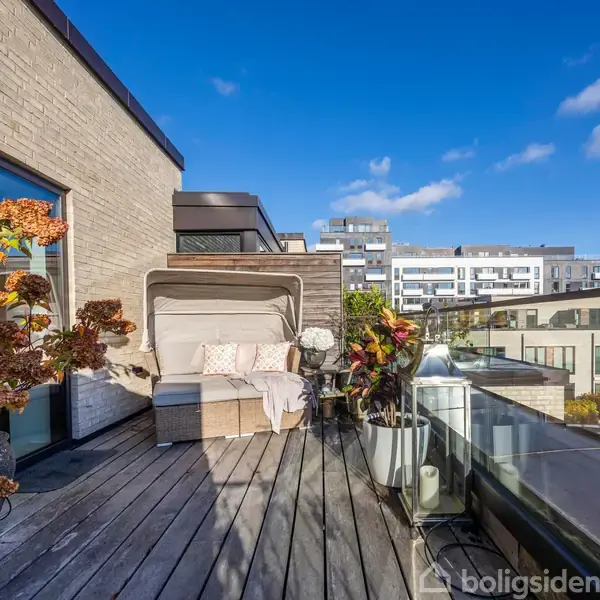 Loungeområde med hvid sofa står stille på en træterrasse. Overdækket med klare skyer og moderne lejligheder i baggrunden. Store planter flankerer siderne af sofaen.