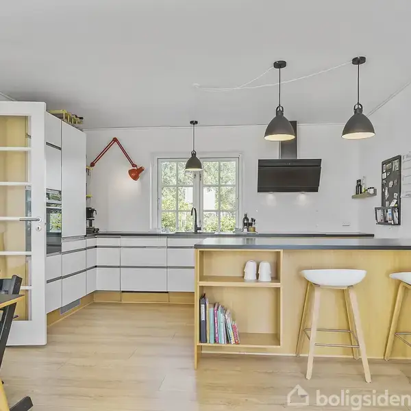 Et lyst køkken med hvide skabe, to barstole ved en køkkenø, hængende pendellamper, og spisestole. Vinduet bagved giver naturligt lys. Et ur og en opslagstavle hænger på væggen.