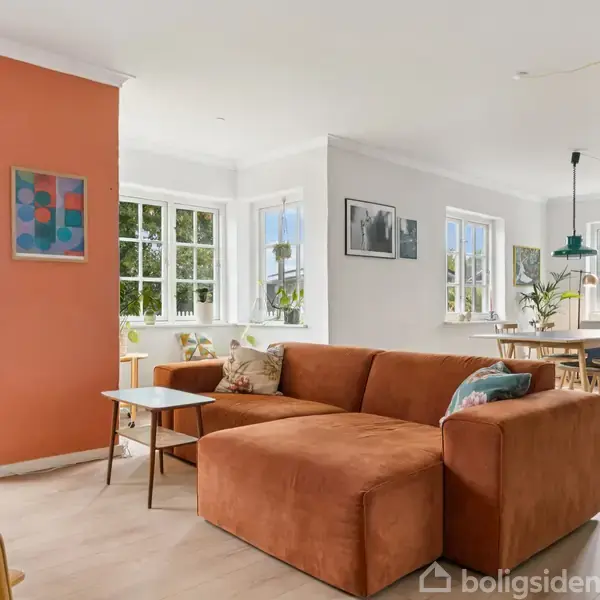 En orange sofa står i et lyst opholdsrum med store vinduer. Til venstre ses en maleriudsmykket væg og en glasdør ud til en have. I baggrunden er et spisebord.