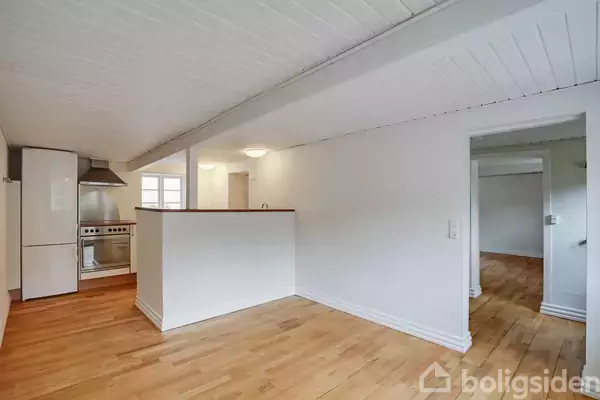 Et lyst køkken med trægulv, hvidmalede vægge og loft. Køkkenet har en komfursektion og et vindue, der giver naturligt lys. En åbning fører videre til et andet rum.