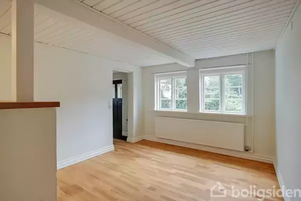 Et lyst rum med trægulv og hvidt loft har tre store vinduer, der lukker naturligt lys ind. I baggrunden er en åben døråbning. Billedet er uden tekst.