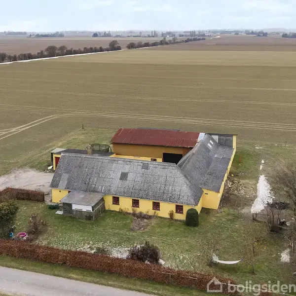Gul landbygning med fladt tag står stille ved en landevej, omringet af marker og lav hæk, i et åbent landskab.