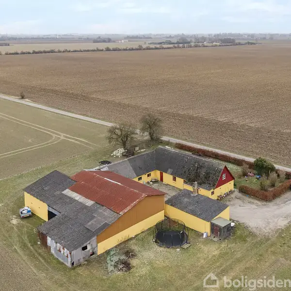 Et gult bondehus med rødt tag og tilhørende bygninger står i en åben marklandskab, omgivet af jord og en lille vej. En trampolin er i gården.