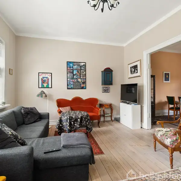 Stue med grå sofa i venstre side, rød sofa og lille bord i midten. Væggene er dekoreret med billeder, og et antikt ur står til højre. Trægulve dominerer rummet.