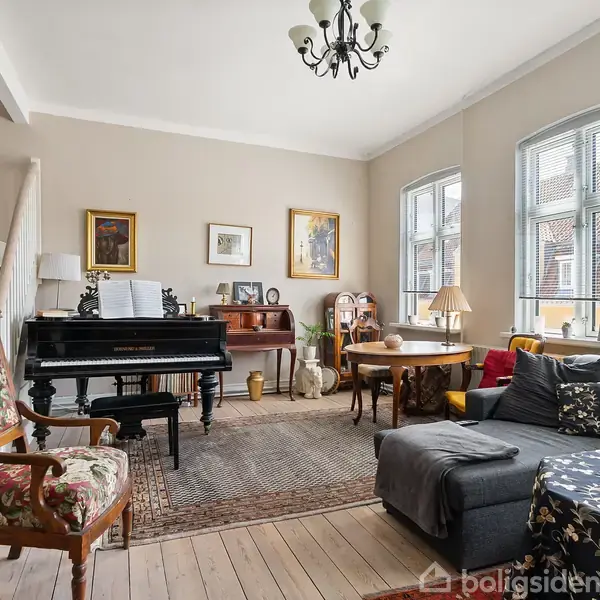 Et klaver står i en stue med diverse møbler, herunder en sofa, lænestol og bord. Væggene er dekoreret med billeder, og der kommer lys ind af tre store vinduer.