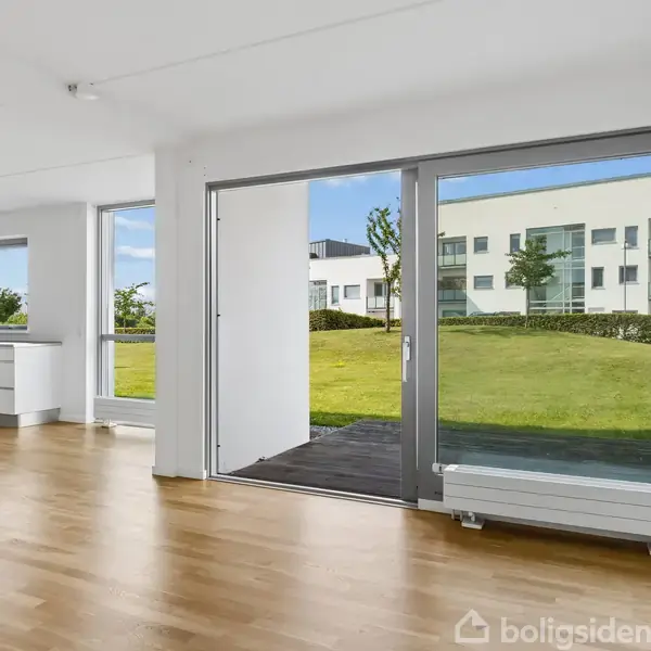Stue med trægulv og store glaspartier, der åbner til en terrasse og udsigt til grønne områder og en moderne bygning. Køkken med hvide skabe til venstre.