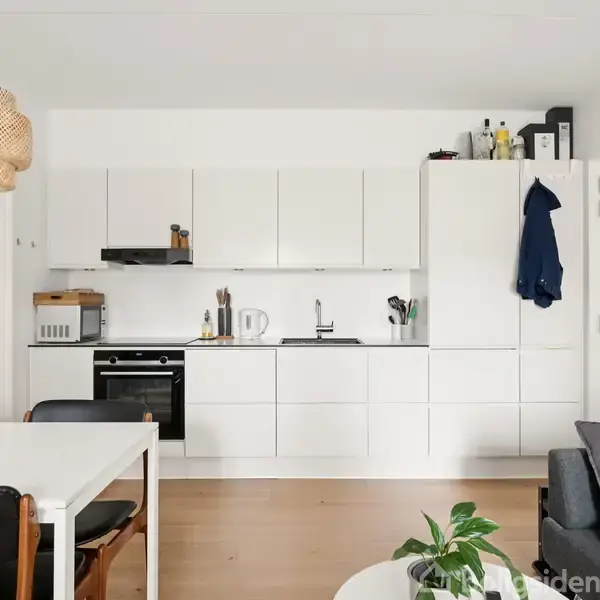 Et moderne køkken med hvide skabe på væggen. En indbygget ovn og diverse køkkenredskaber er synlige. Ved siden af står et spisebord med stole. Hængende lampe over bordet. Ingen tekst fundet.