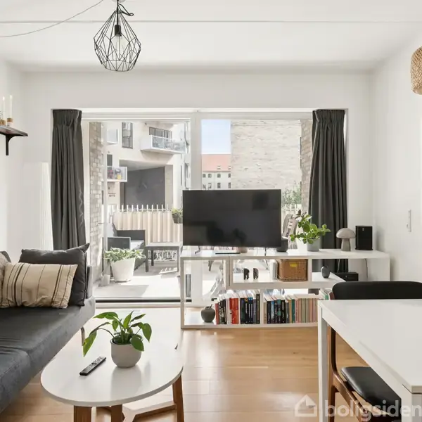 Stue med en sofa, der står ved siden af et sofabord med en plante på. Et TV står på et bord foran vinduer med sorte gardiner. Der er en spisebord i nærheden.