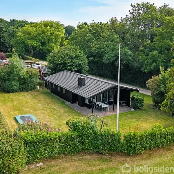Et sort hus står stille på en velplejet græsplæne, omgivet af buske og træer. En flagstang står ved siden af huset, og en trampolin er placeret på plænen.