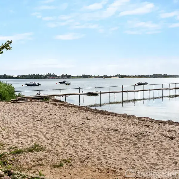 Sandstrand med en lang, smal bådebro strækker sig ud over stille vand. Både flyder roligt på søen. Grønt buskads og træer omgiver området, under en let overskyet himmel.
