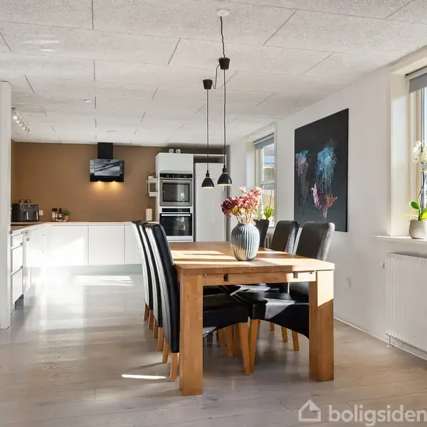 Spisebord med sorte stole står i solbelyst køkken-alrum. Væggene prydes med kunst, og store vinduer slipper lys ind, mens planter og blomster dekorerer rummet med naturlig grønne nuancer.