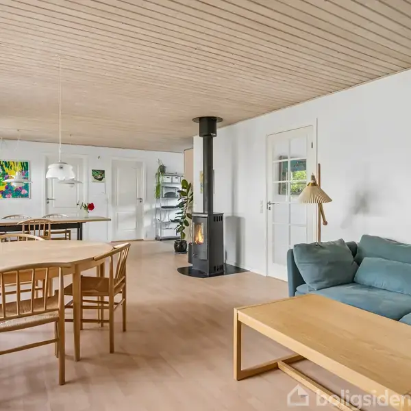 Stue med spisebord, stole og en blå sofa. Brændeovn står ved en hvid væg med hængende kunst. Trægulv og lyst trægulv, grøn plante nær spiseplads. Væggene har farverige malerier.