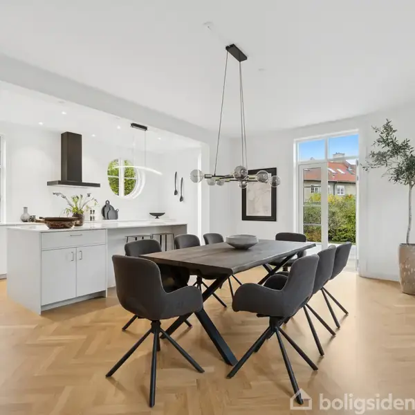 Et moderne køkken-alrum med et langt spisebord og seks stole. Rummet er lyst med store vinduer, en lysekrone over bordet, og et oliventræ i hjørnet.