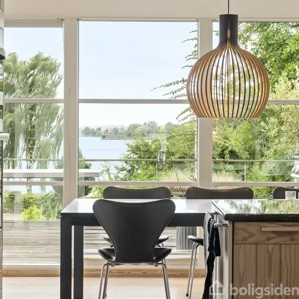 Køkkenbord med stole står mod store vinduer, der åbner ud til en veranda med udsigt over en sø og træer. En moderne pendellampe hænger over bordet.