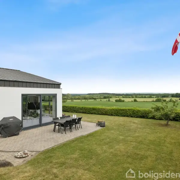 Et moderne hus med store glasdører åbner ud mod en terrasse, hvor der står et bord med stole. Dannebrog vajer fra en flagstang i en velplejet have med udsigt over marker.