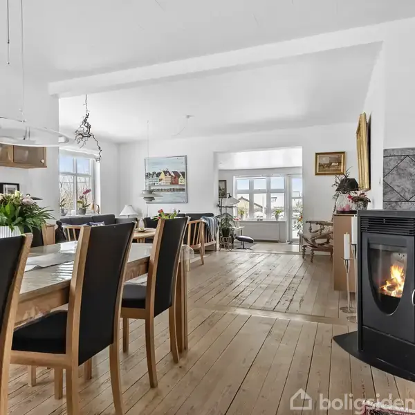 Et spisebord med sorte stole står i et lyst rum. En tændt pejs opvarmer området, mens naturligt lys strømmer ind gennem store vinduer. Blomster pynter bordet.