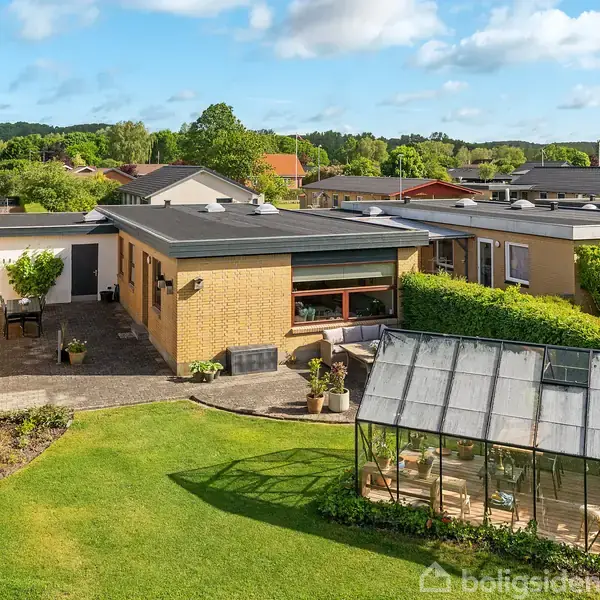 Et enplanshus med fladt tag omgivet af en velplejet have. En terrasse med borde og stole ligger ved huset. Et drivhus står på græsplænen. Solskin og nabohuse i baggrunden.