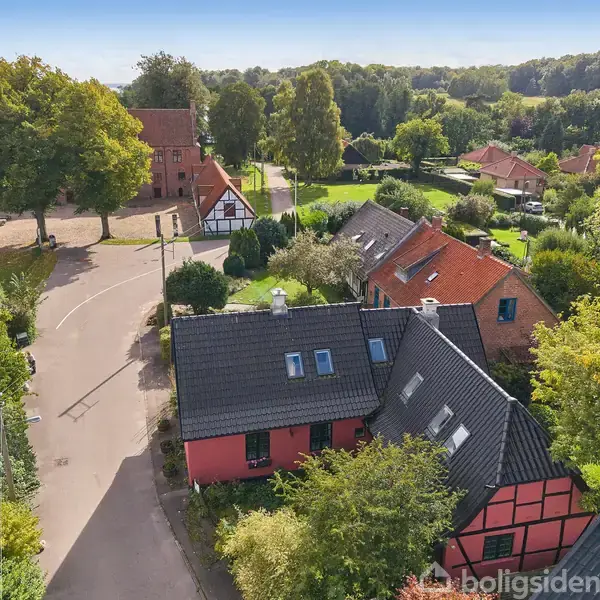 Røde bindingsværkshuse står stille i en solrig landsby med grønne træer, haver og snoede veje i baggrunden, hvilket skaber en landidylisk stemning.