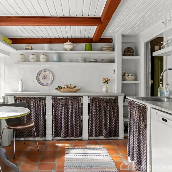 Køkken med hvidt loft og røde bjælker. Rundt bord med to stole, åbne hylder med tallerkener og krukker. Mørke stofskørter skjuler opbevaring under bordpladerne, tæppe på terracotta-gulv.