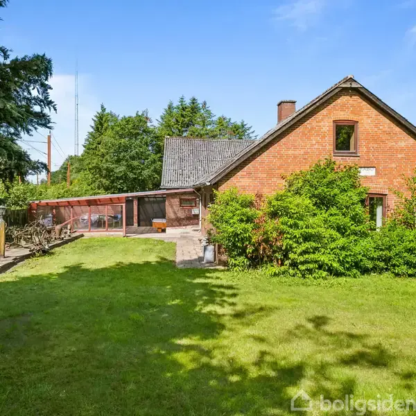 En rød murstensbygning med græsplæne foran, omgivet af buske og træer. Det er solrigt, og der er et overdækket område til venstre ved siden af huset.