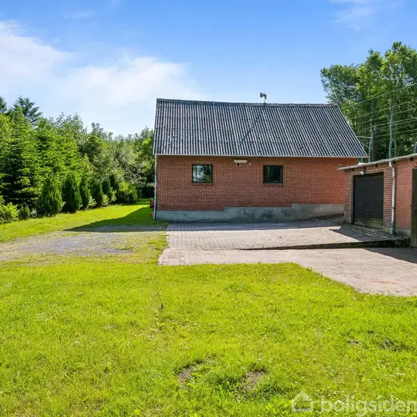 Et rødt murstenshus står stille midt i en have med græs og træer langs siden. En flisebelagt indkørsel fører op til huset med blå himmel over.