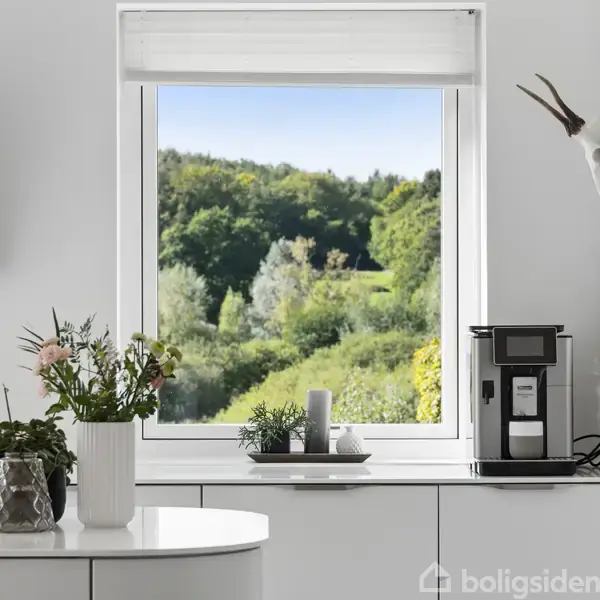 En moderne køkkenindretning med en blomsterbuket og espressomaskine på en hvid bordplade. Gennem vinduet ses frodig grønt landskab udenfor. En vægdekoration hænger til højre.