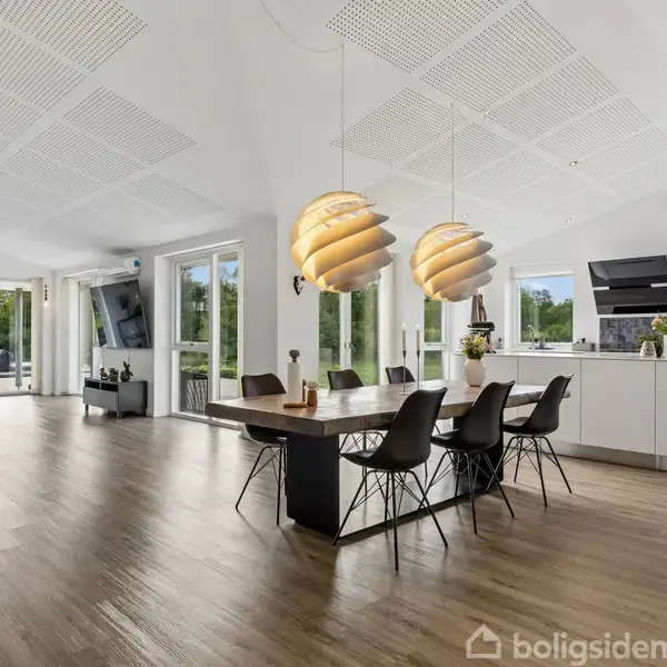 En rummelig, moderne stue-køkken kombination. Et stort spisebord med sorte stole står centralt, omgivet af minimalistiske møbler og store vinduer, der giver udsigt til grønne omgivelser udenfor.