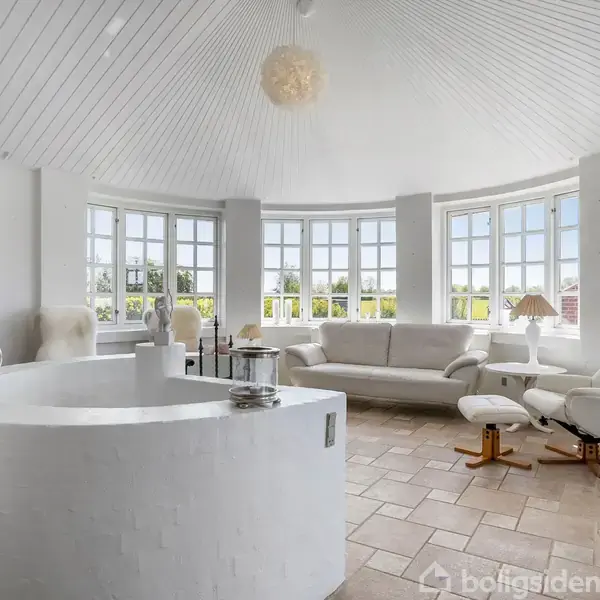 Hvidt, lys stue med en rund sofa, lænestole og et sofabord. Store vinduer omslutter rummet og giver udsigt til det grønne udenfor. En maleri pryder væggen.