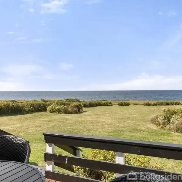 Terrasse med sorte stole og bord med udsigt over en grøn græsplæne, buske og horisonten af havet under en blå himmel.