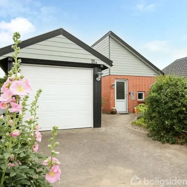 Garagen med hvid port står ved et rødt murstenshus. Rosa blomster vokser i forgrunden, mens grønne buske flankerer den brolagte indkørsel. Nummeret "11 A" ses på garagen.