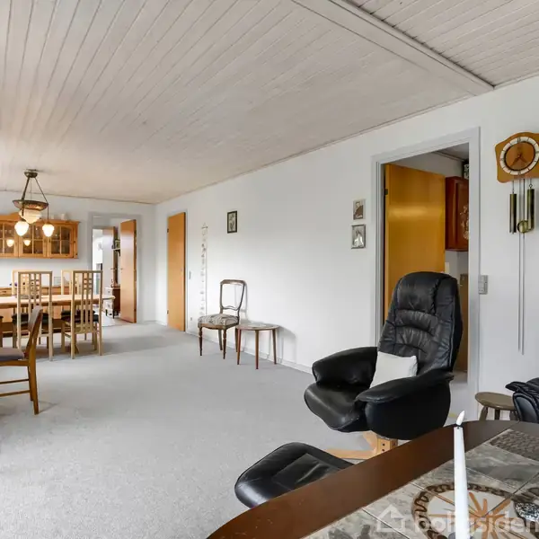 Stue med lænestole og et sofabord i forgrunden. I baggrunden er der et spisebord med stole under en hængende lampe. Væggene er hvide med en træloft.