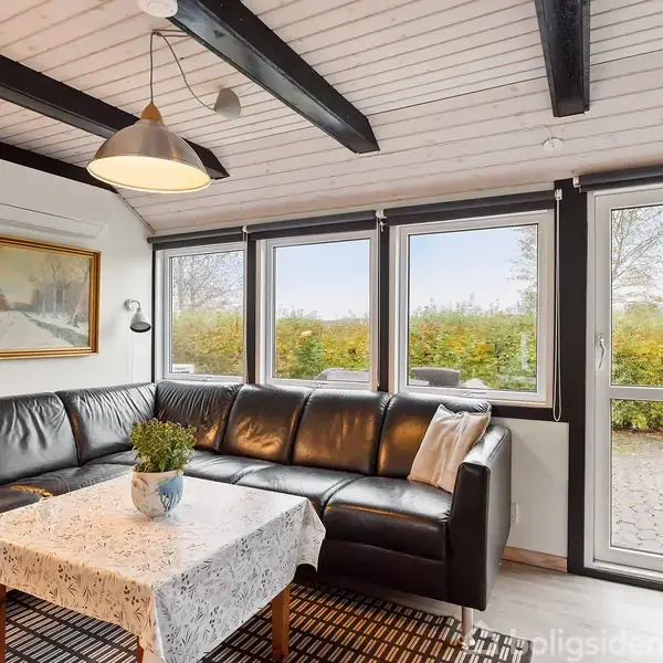 Sort lædersofa står i en stue, omgivet af store vinduer med udsigt til en hæk. Et maleri hænger på væggen, og et bord med dug og plante står foran sofaen.