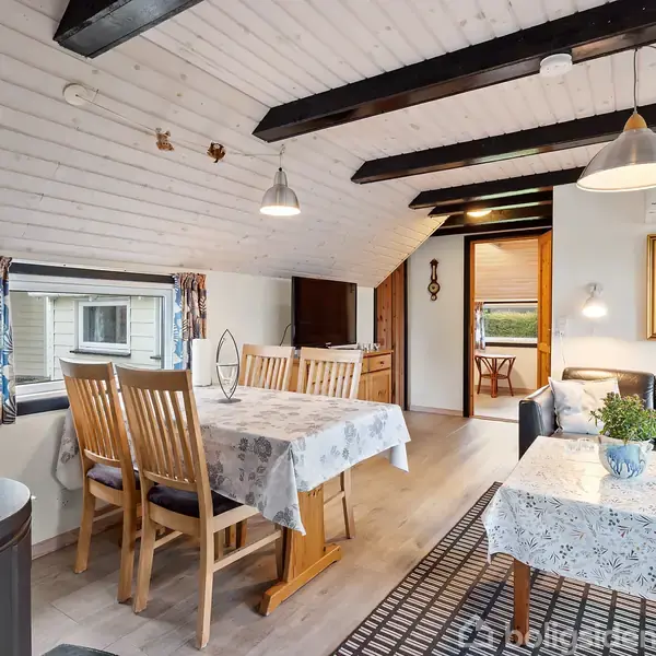 Spisestue med spisebord dækket med dug, træstole med hynder, sofa, sofabord, vægmaleri, skråt loft med bjælker og vindue med gardiner. Hængende lamper giver belysning.